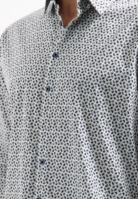 Shirt in lichte stof met een geometrisch patroon van kleine marineblauwe driehoeken op een lichte achtergrond; heeft een kraag en blauwe knopen.