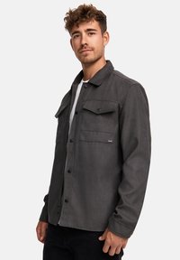 Chaqueta gris de botonadura con dos bolsillos en el pecho, hecha de una tela suave. Presenta un cuello con solapa y un corte recto. Lleva puesta sobre una camisa blanca.