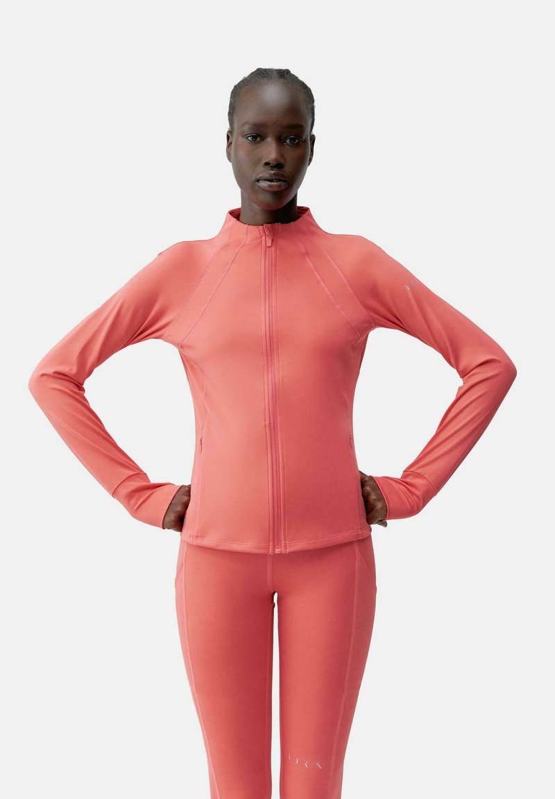 Fittad atletisk jacka i ljus korall, tillverkad av ett slätt, stretchigt tyg. Har hög krage, långa ärmar och hel dragkedja framtill.