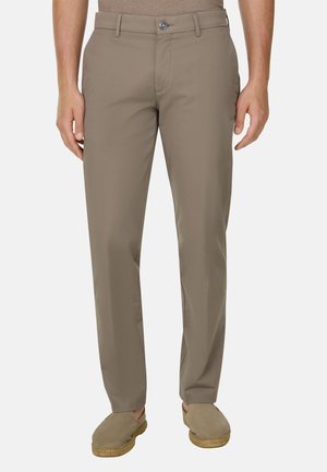 Mężczyzna noszący beżowe spodnie o prostych nogawkach oraz dopasowane wsuwane buty, stojący na tle jednolitego jasnego tła.