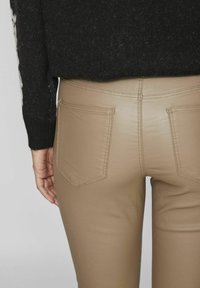 Bež hlače iz umetnega usnja s tesnim krojem, zadnjimi žepi in subtilnimi šivi. Nosi se z črnim teksturiranim puloverjem.