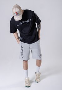 Black short-sleeve T-shirt with white logo, gray shorts with black lettering, beige cap with graphic, white socks, and beige sneakers.