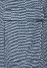 Blå tyg med fiskbensmönster och en platt ficka. Materialet verkar ha en textur och har ett subtilt diagonalt randmönster.