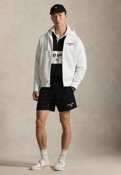 Sweat à capuche blanc zippé porté sur une chemise noire à col avec un logo rouge et blanc, associé à un short noir et des baskets blanches. Accentué par des chaussettes côtelées.