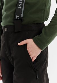 Zwarte outdoorbroek met ritszak, gecombineerd met een groene fleece top. Voorzien van verstelbare zwarte bretels met witte letters.