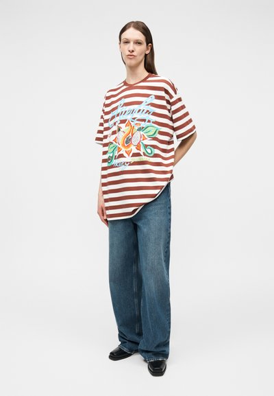 Άτομο που φοράει καφέ και λευκό ριγέ φαρδιά μπλούζα με λουλουδάτο σχέδιο και μπλε φαρδιά τζιν, στέκεται με τα χέρια πίσω από την πλάτη.