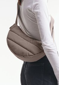Sac bandoulière rembourré marron avec une silhouette courbée, des motifs de piqûres diagonales, une fermeture zippée et une sangle réglable. Logo visible sur le devant.