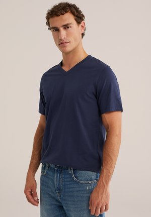 Homme aux cheveux bouclés portant un t-shirt bleu marine à col en V et un jean bleu, debout devant un fond clair uni.