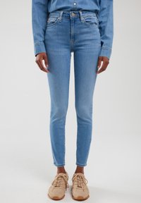 Žena u svijetloplavim trapericama s visokim strukom, traper košulji i bež tenisicama stoji na bijeloj pozadini.