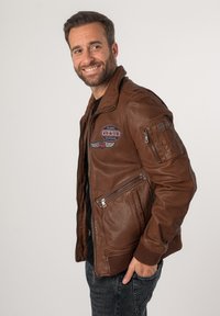 Braune Lederbomberjacke mit gerippten Bündchen; verfügt über gestickte Aufnäher und Reißverschlusstaschen; glatte Textur und taillierte Passform.