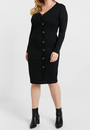 Kobieta ubrana w czarną, zapinaną na guziki sukienkę z długim rękawem sięgającą kolan, w zestawieniu z czarnymi botkami za kostkę, stojąca na tle jasnego, jednolitego tła.