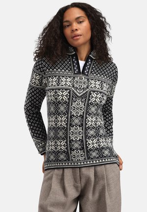 Kvinne med krøllete hår som har på seg en svart- og hvitmønstret strikkegenser med høy krage og grå vidbukser, hendene i lommene.