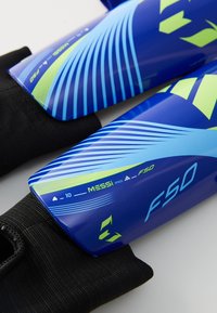 Mėlynos ir žalios Adidas F50 Messi futbolo šinų apsaugos, laikomos ant juodų apsauginių rankovių.