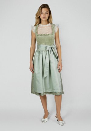 Junge Frau in einem salbeigrünen traditionellen Kleid mit einem floralen, transparenten Oberteil und gebundener Satin-Schürze, stehend vor einem einfarbigen grauen Hintergrund.