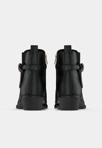 Melnas potītes zābaki ar sānu sprādzēm un zemu blokveida papēdi, attēloti no aizmugures uz vienkārša baltā fona.