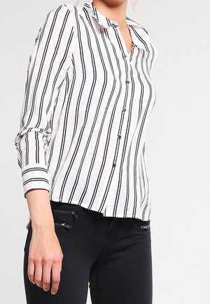 Vrouw draagt een wit shirt met lange mouwen met zwarte verticale strepen en een zwarte broek met ritsdetails op de zakken.