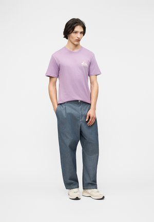 Mladý muž stojaci s rukami v kapsách, oblečený v lila tričku s potlačou, voľné modré džínsy a biele tenisky na jednoduchom pozadí.