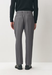 Pantalon gris à coupe ajustée, avec poches arrière, passants de ceinture et une texture lisse. Porté avec des chaussures noires.