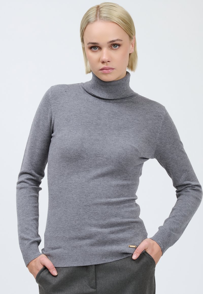 Pull en col roulé gris en tissu tricot doux, avec des manches longues et un ourlet côtelé. Petit logo métallique sur le coin inférieur gauche.