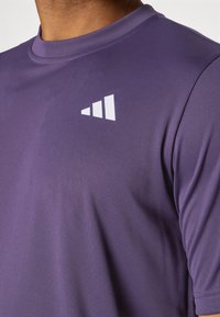 Purple athletic shirt with a smooth texture, featuring a white logo in a triangular shape on the upper left chest area. Short sleeves.