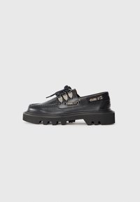 Chaussures en cuir noir avec une semelle en caoutchouc épaisse, présentant des coutures proéminentes et des accents décoratifs en métal sur les côtés.