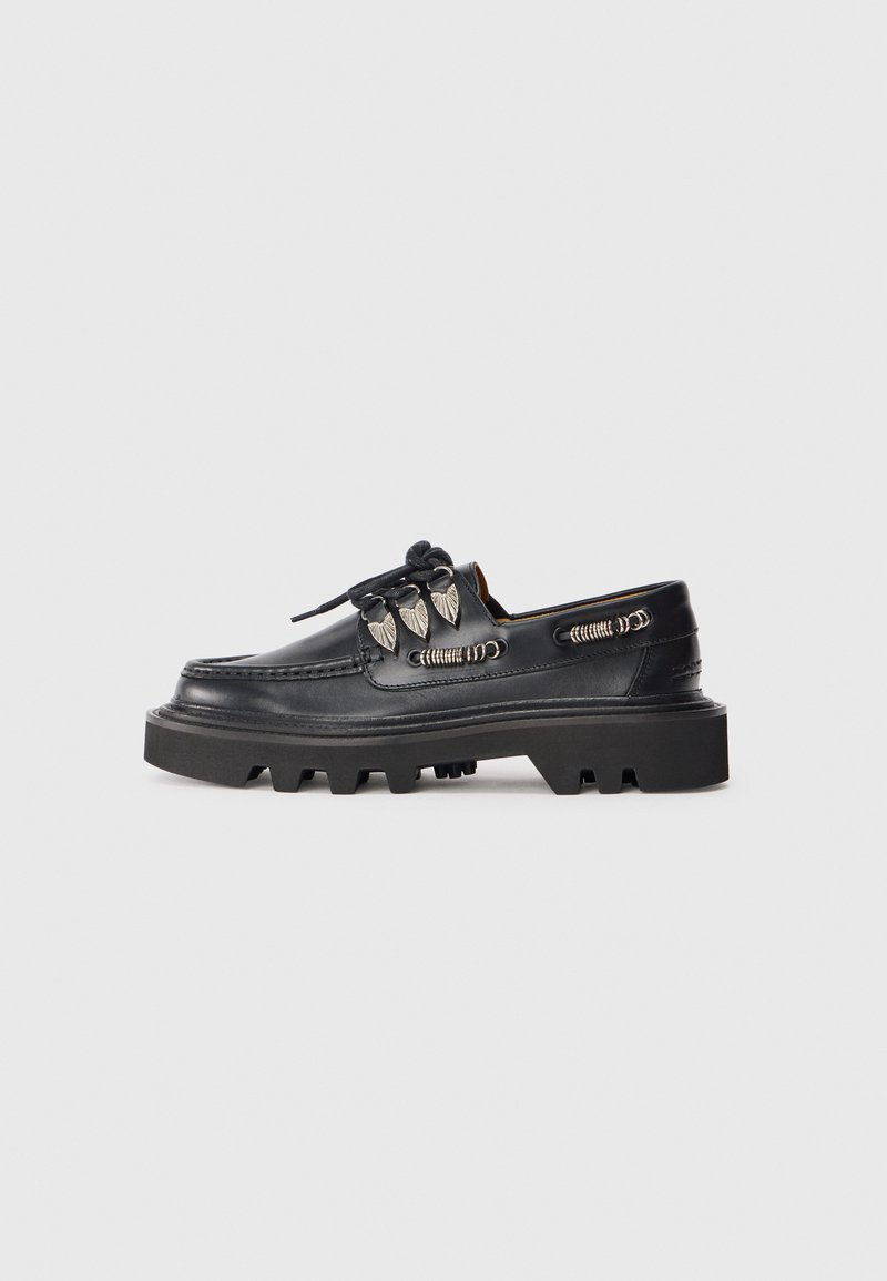 Chaussures en cuir noir avec une semelle en caoutchouc épaisse, présentant des coutures proéminentes et des accents décoratifs en métal sur les côtés.