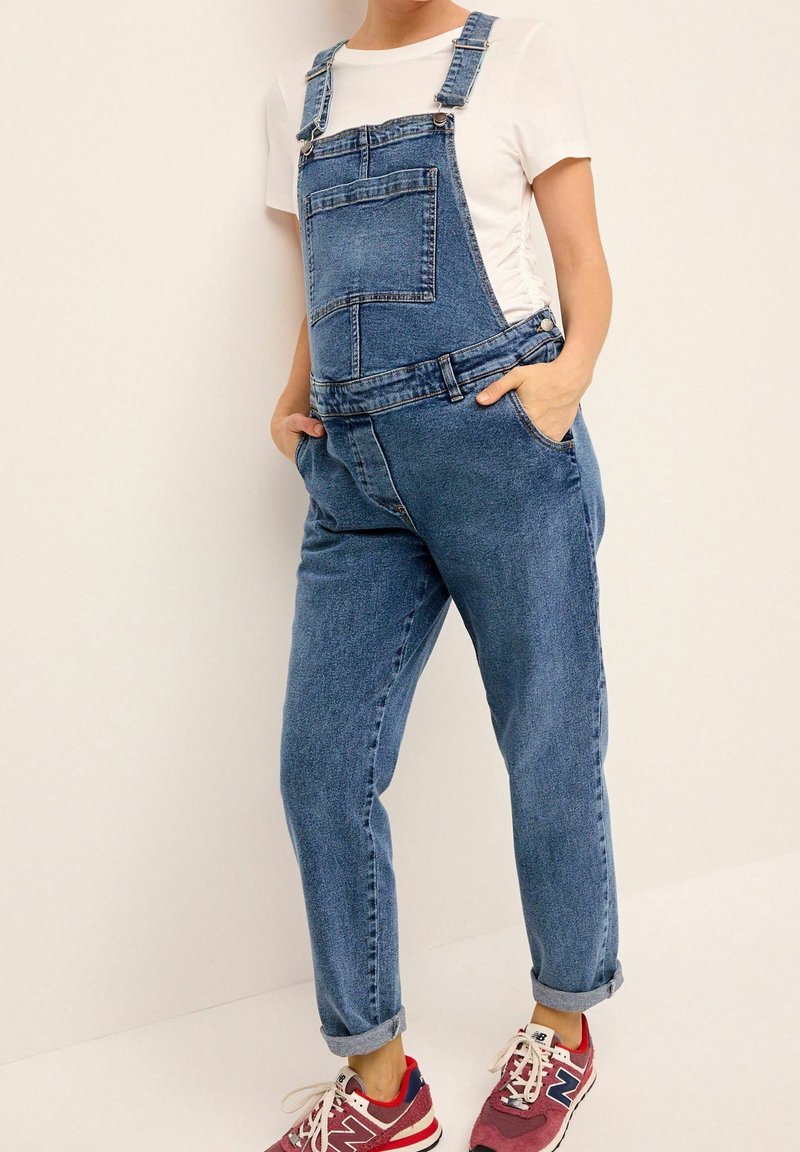 Denim tuinbroeken in middelblauw met verstelbare banden, een voorkantzak en taps toelopende benen. Gecombineerd met een wit t-shirt en rode sportschoenen.