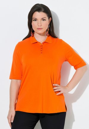 Kobieta o długich ciemnych włosach, ubrana w pomarańczową, krótką koszulkę polo i kolorowe, wiszące kolczyki, stoi z jedną ręką na biodrze.