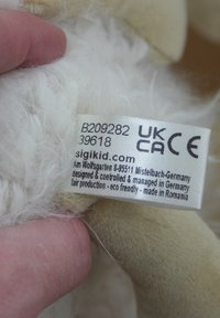 Stuffed animal tag with white fabric and embossed text. Features soft white fur and beige accents. Includes production details and website.