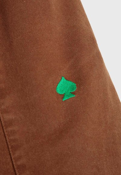 Un tejido marrón con una textura suave presenta un logo de trébol bordado en verde, centrado en la parte inferior izquierda.