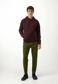 Sweat à capuche bordeaux avec cordon de serrage et poches zippées noires, assorti à un pantalon de jogging vert et des chaussures de sport noires.