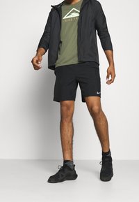 Svart lättviktig jacka över en grön t-shirt, svarta shorts och svarta träningsskor. Funktioner inkluderar en Nike-logotyp och randiga strumpor.