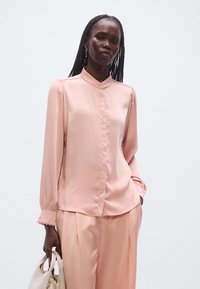 Lyserød satinbluse med mandarin-krave, knaplukning og detaljer med rynkede skuldre. Kombineret med matchende bleg lyserøde bukser og en håndtaske.