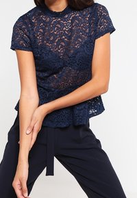 Marinblå spetsblus med korta ärmar, scallopad halsringning, intrikata blommönster och en avslappnad silhuett, ihop med mörka byxor.