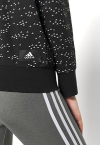 Sudadera deportiva negra con un patrón triangular blanco, que presenta un parche con el logotipo negro de Adidas. Combinada con leggings grises con rayas blancas.
