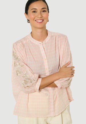 Vrouw glimlacht met armen over elkaar, draagt een lichtroze geruite blouse met gouden bloemrijke borduursels op de mouwen en beige broek.