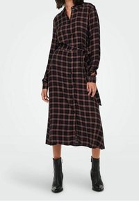 Frau trägt ein langärmliges, schwarz-rotes karierter Midikleid mit Taillengurt und schwarzen Knöchelstiefeln, stehend vor einem weißen Hintergrund.