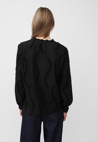 Camicia nera con dettagli a rouches testurizzati in un motivo ondulato, maniche lunghe e scollatura leggermente arricciata. Indossata con jeans scuri.