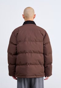 Chaqueta acolchada marrón con un cuello de pana negro, con amplias costuras horizontales y un ajuste relajado, llevada sobre pantalones grises.