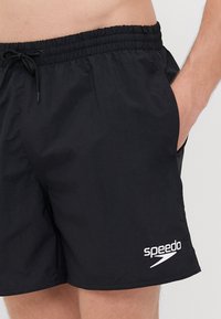Juodi maudymosi šortai su elastinga juostos juosta, raišteliu, šoninėmis kišenėmis ir baltu "Speedo" logotipu kairėje kojoje.