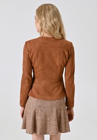 Blonde vrouw in een bruine suède jas en een gestructureerde bruine rok, staand met haar rug naar de camera tegen een eenvoudige witte achtergrond.