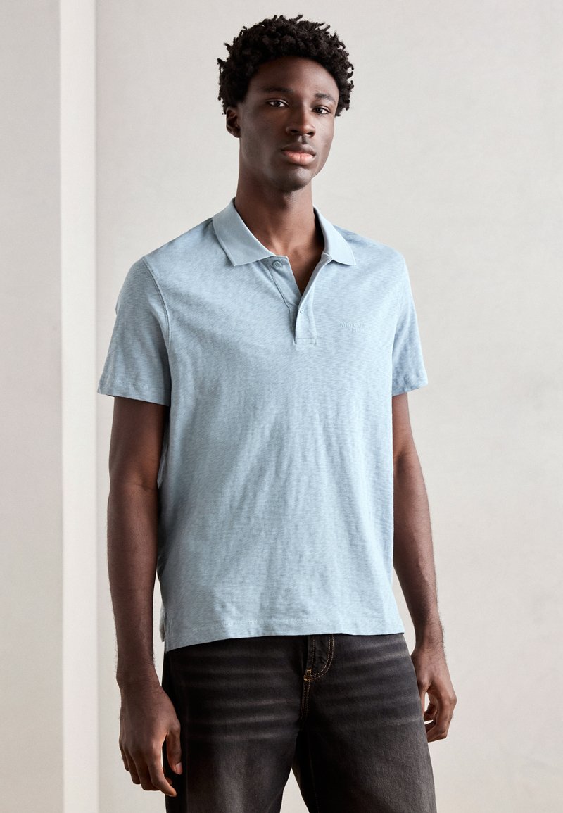 Polo shirt azzurro chiaro realizzato in un tessuto morbido, con colletto classico, maniche corte e una leggerissima trama melangiata. Abbinato a jeans scuri.