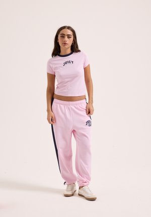 Woman wearing light pink Juicy brand t-shirt and matching sweatpants with side stripes, standing against a plain light background.