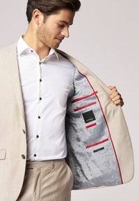 Beige Sakko mit einem hellgrauen gemusterten Innenfutter und roten Akzenten, Knopfverschluss und zwei Innentaschen. Weißes Hemd mit braunen Knöpfen.