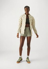 Lys beige dunjakke med glidelåslukking, ribbet tekstur og oransje detaljer. Kombinert med grønne shorts og grønne terrengsko.