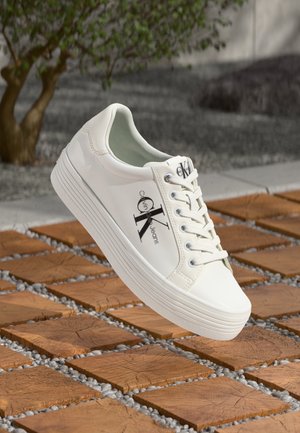 Biały sneaker Calvin Klein z sznurowadłami i logo z boku, unoszący się nad drewnianą podłogą z małymi szarymi kamieniami między płytkami.