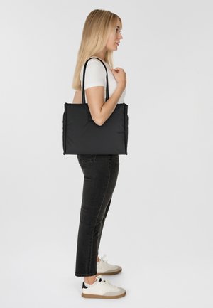 Zwarte schoudertas met een strak ontwerp, stoffen textuur en lange hengsels, gedragen door een vrouw in een witte blouse en zwarte spijkerbroek.
