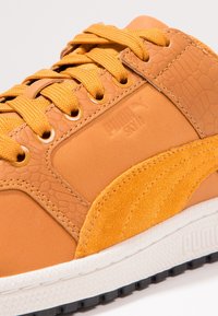 Baskets Puma en cuir synthétique orange avec des accents texturés, des détails perforés, et une semelle en caoutchouc blanche avec des crampons noirs.