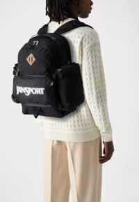 Zwarte Jansport rugzak met meerdere ritssluitingen, stoffen label, verstelbare banden en een gestructureerd ontwerp. Gekozen over een witte trui.