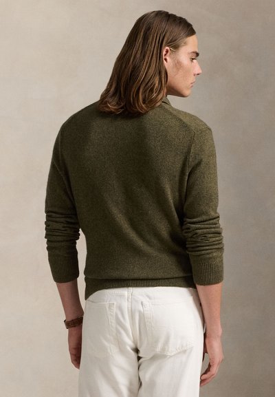 Mies, jolla on olkapäille ulottuvat hiukset, yllään oliivinvihreä neule ja valkoiset housut, selin yksinkertaista beigeä taustaa vasten.